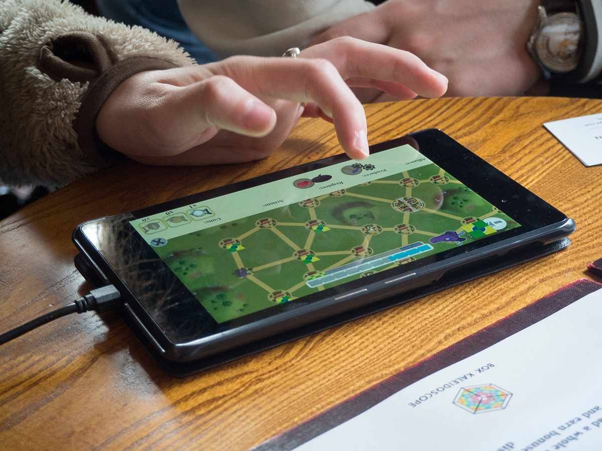 The game running on a tablet device. A player's index finger is poised above one of the settlements as they consider their next move.